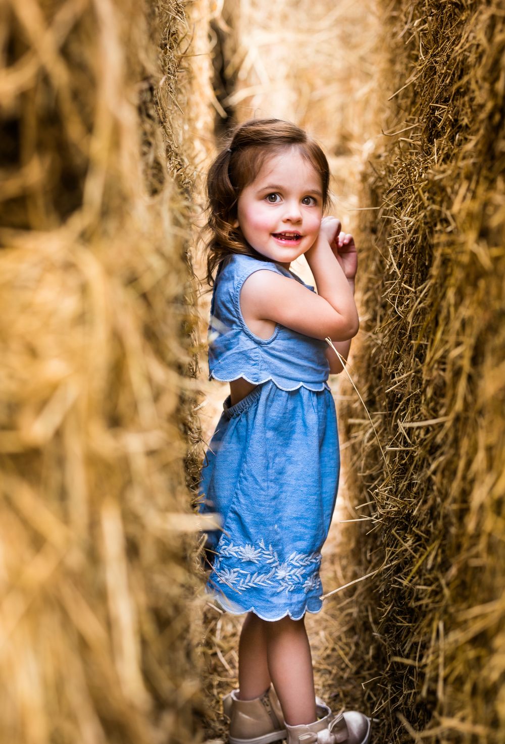 Young child in blue dress peeking through hay bales in warm lighting.