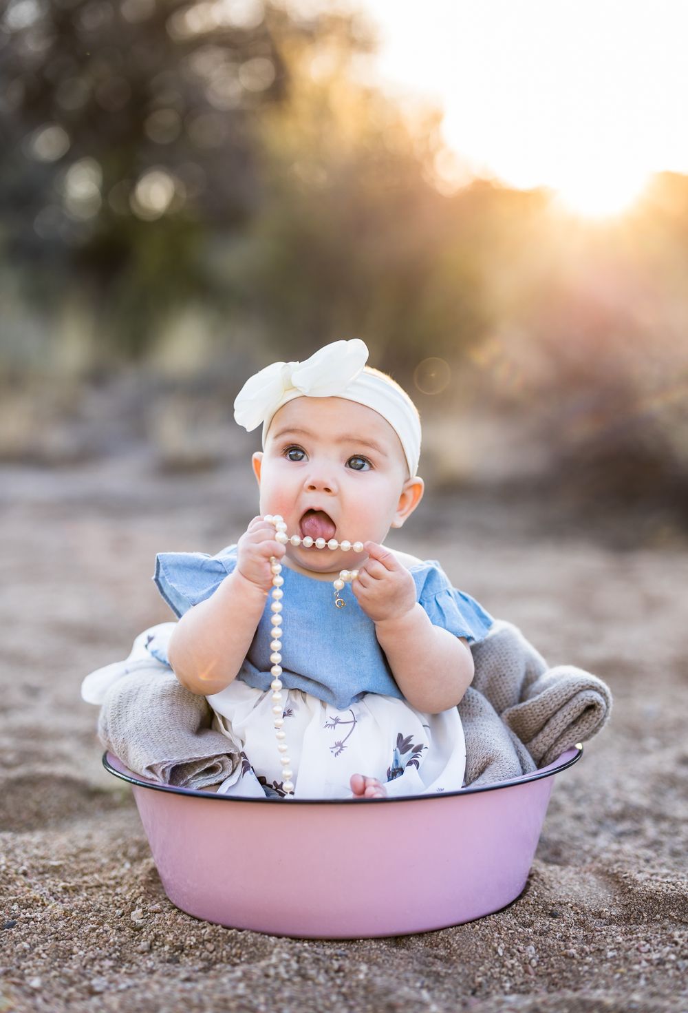 A baby in blue clothing sits in a pink wash basin outdoors at sunset.