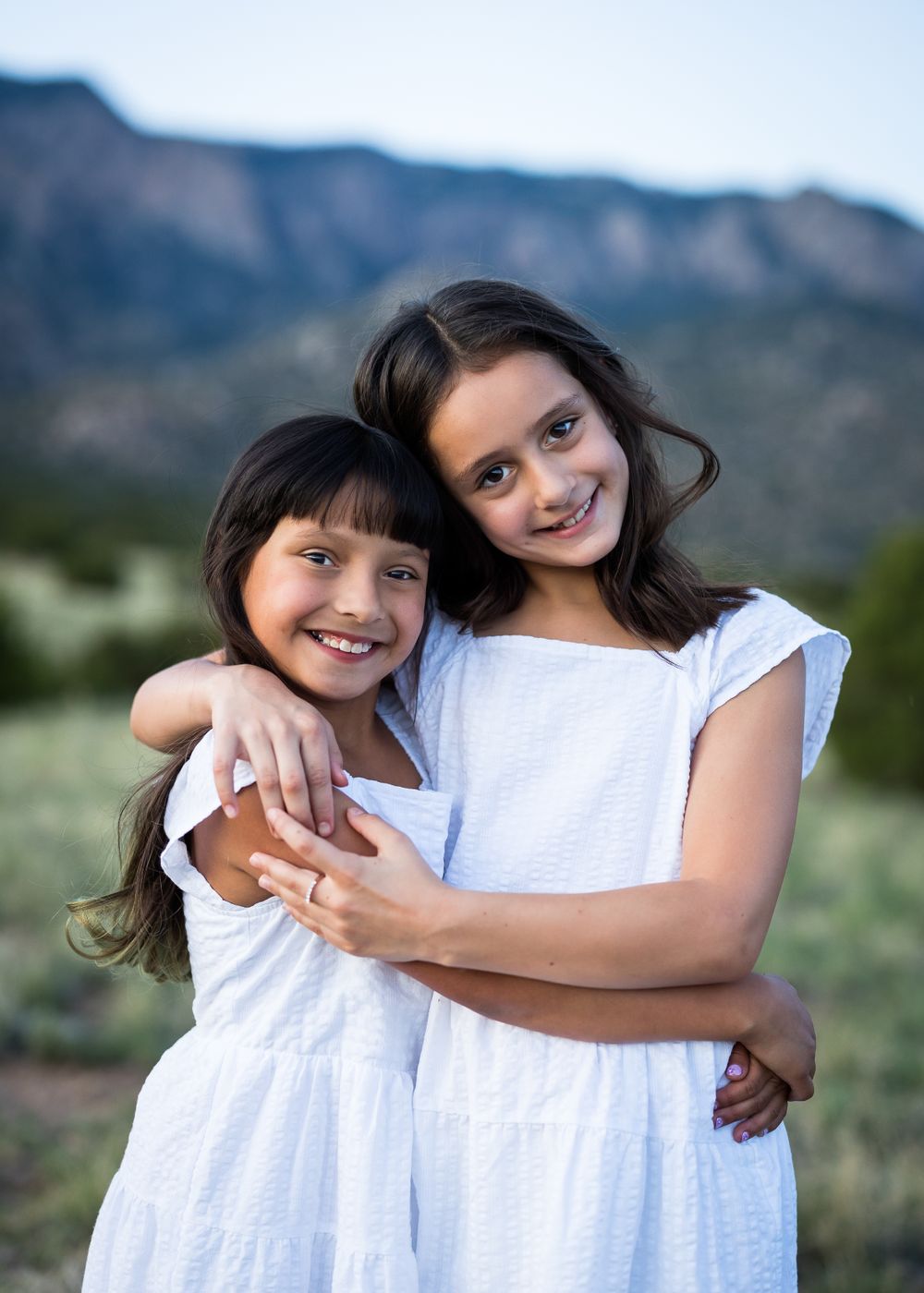 Two children in white dresses embracing against a mountainous backdrop.