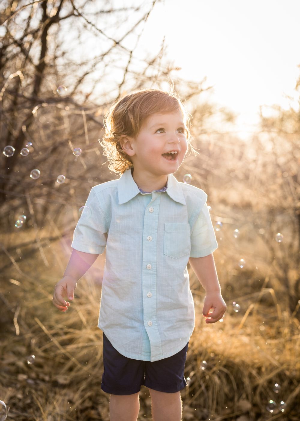 Child plays with bubbles in sunlit nature setting wearing light blue shirt.