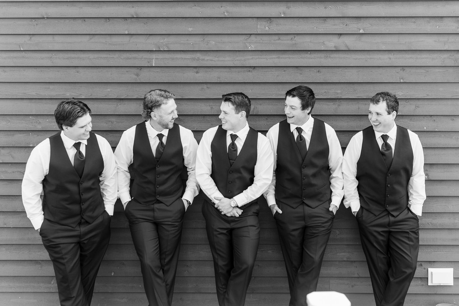 Wedding party in dark suits poses against gray wooden wall in black and white.