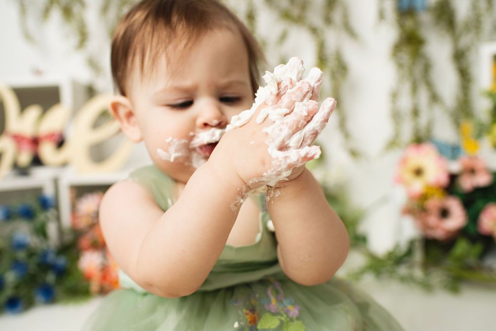Cute baby girl in green dress enjoying her first birthday cake smash with frosting on her hands and face.