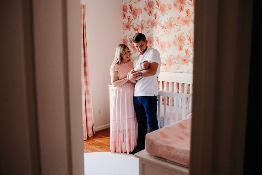 New parents in pink nursery tenderly holding their newborn baby by a white crib.