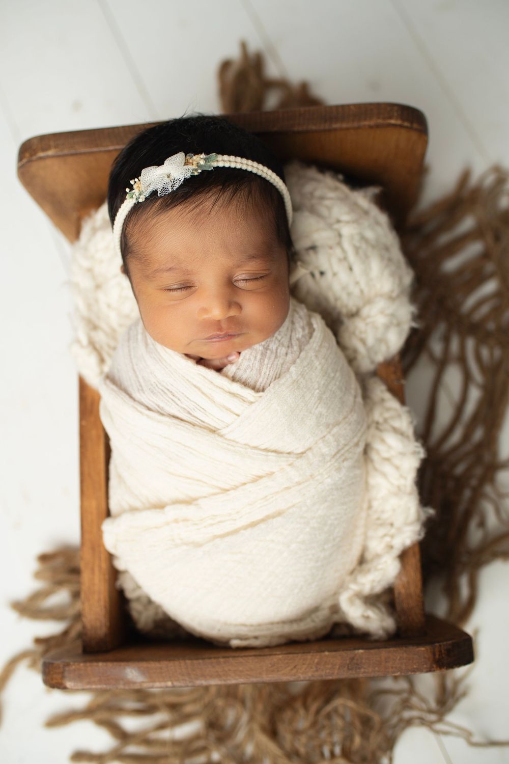 Peaceful newborn baby girl swaddled in cream wrap with pearl headband sleeping in rustic wooden bed.