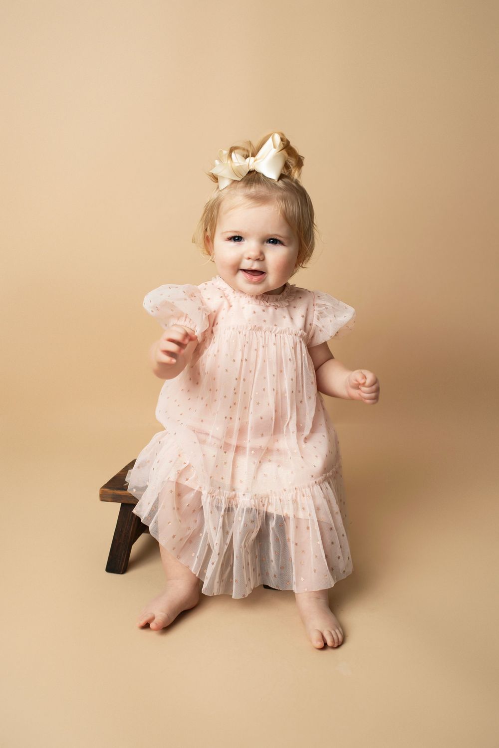 Adorable toddler girl in pink tulle dress and gold crown sitting on a small wooden stool.