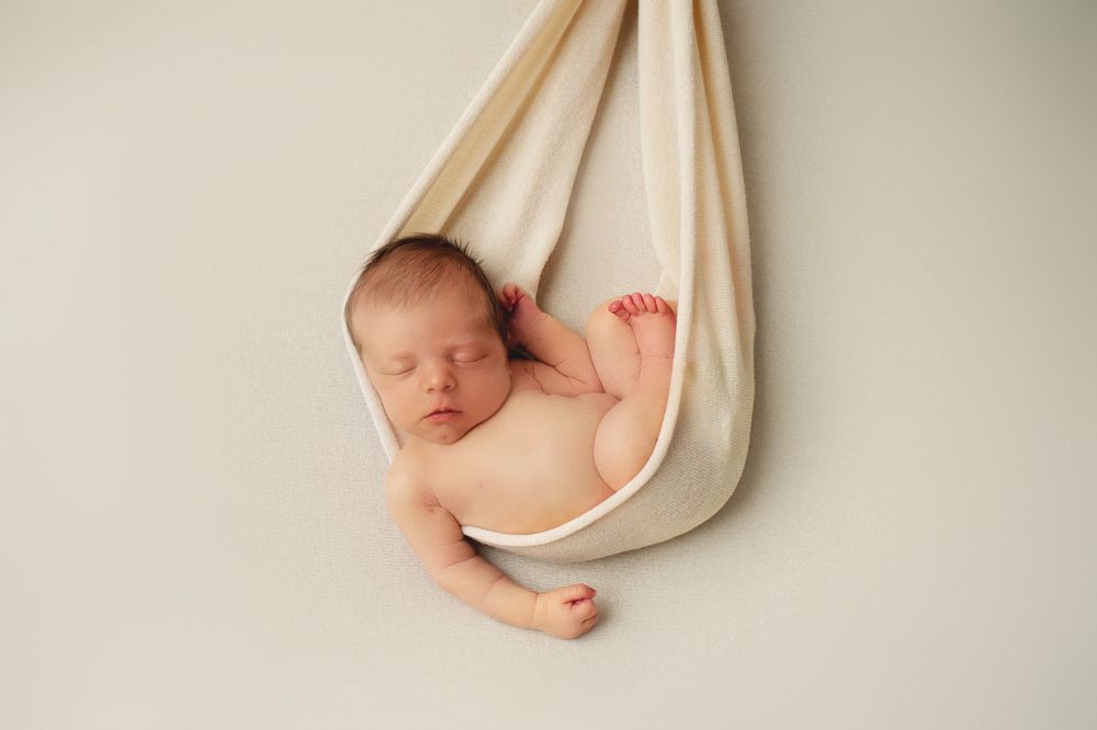 Sleeping newborn baby posed in cream fabric hammock swing on neutral white background.