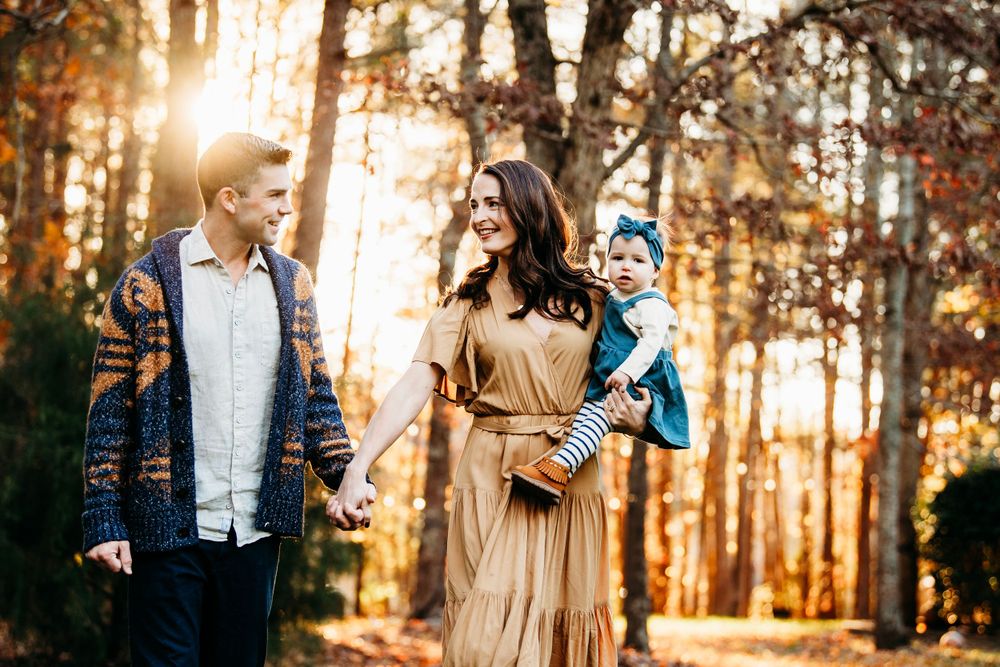 Happy young family with baby walking through golden autumn forest in warm fall clothing.