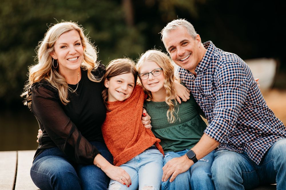 Happy family of four with two daughters smiling together outdoors near a sunlit lake.