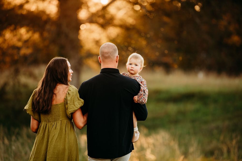 Family of three with toddler standing in golden sunset meadow viewed from behind.