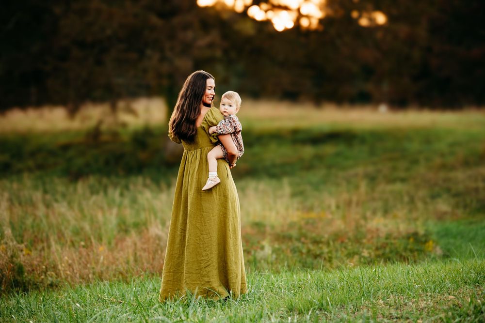 Mother in olive green dress holding her baby in a sunlit autumn field at sunset.