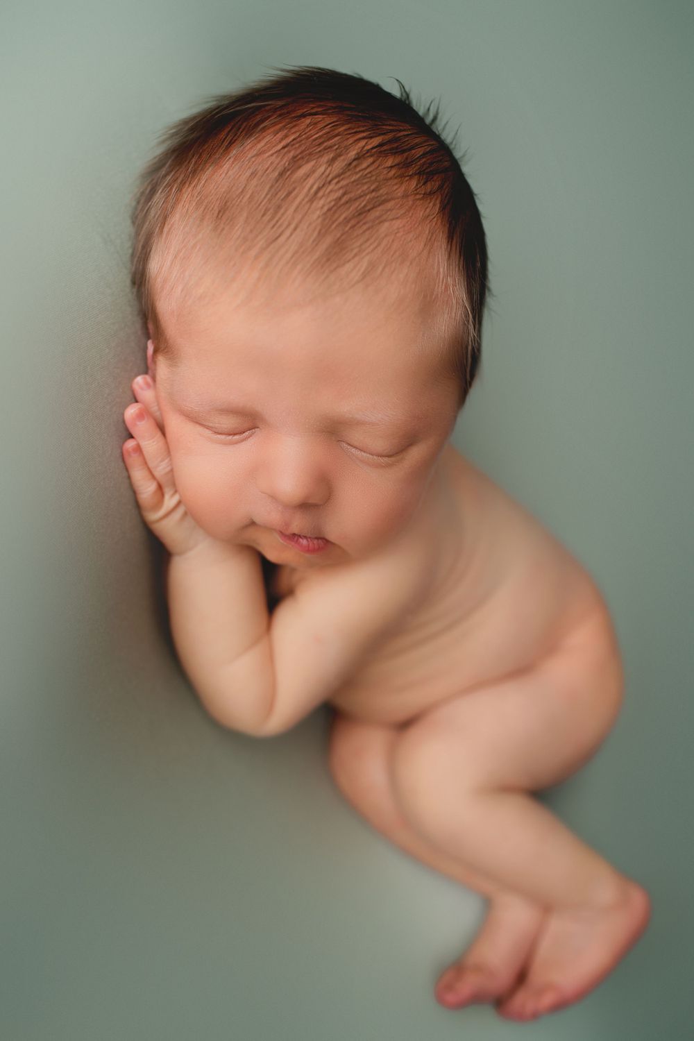 Peaceful newborn baby with dark hair sleeping curled up on soft mint green background.