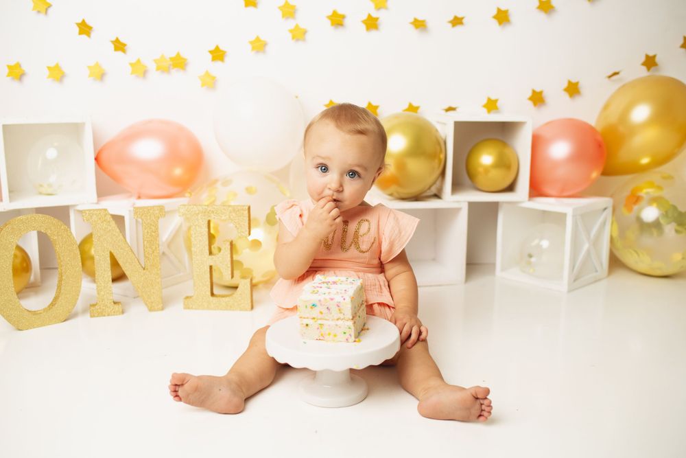 Baby girl in pink dress celebrates first birthday smashing cake surrounded by gold balloons and ONE decorations.