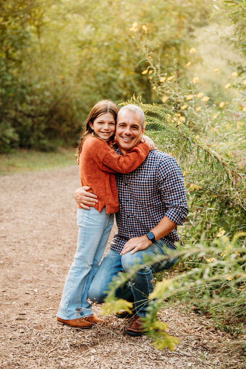 Smiling father kneeling on nature path while daughter in orange sweater hugs him outdoors.