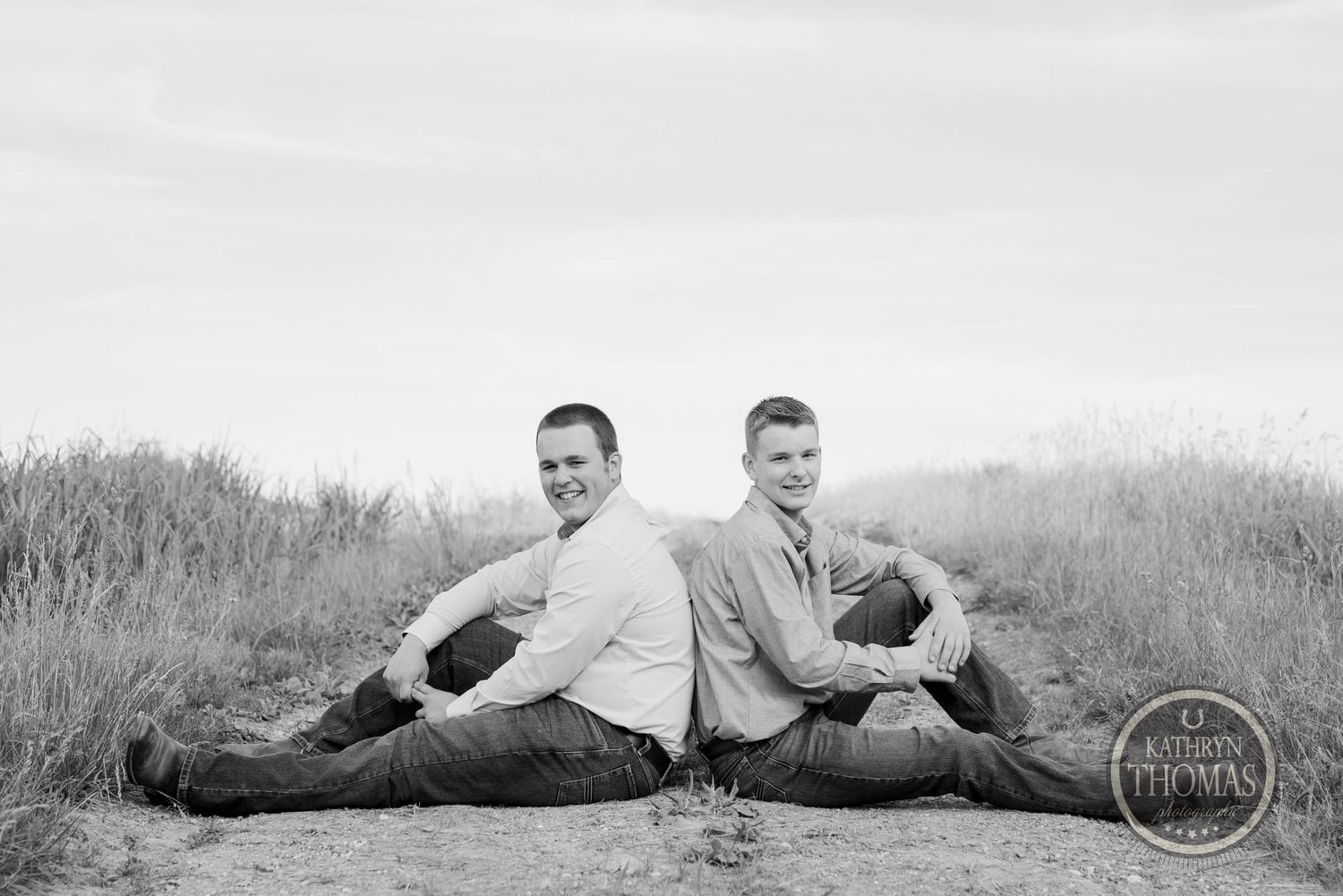 Black and white portrait of two people sitting back to back in field.