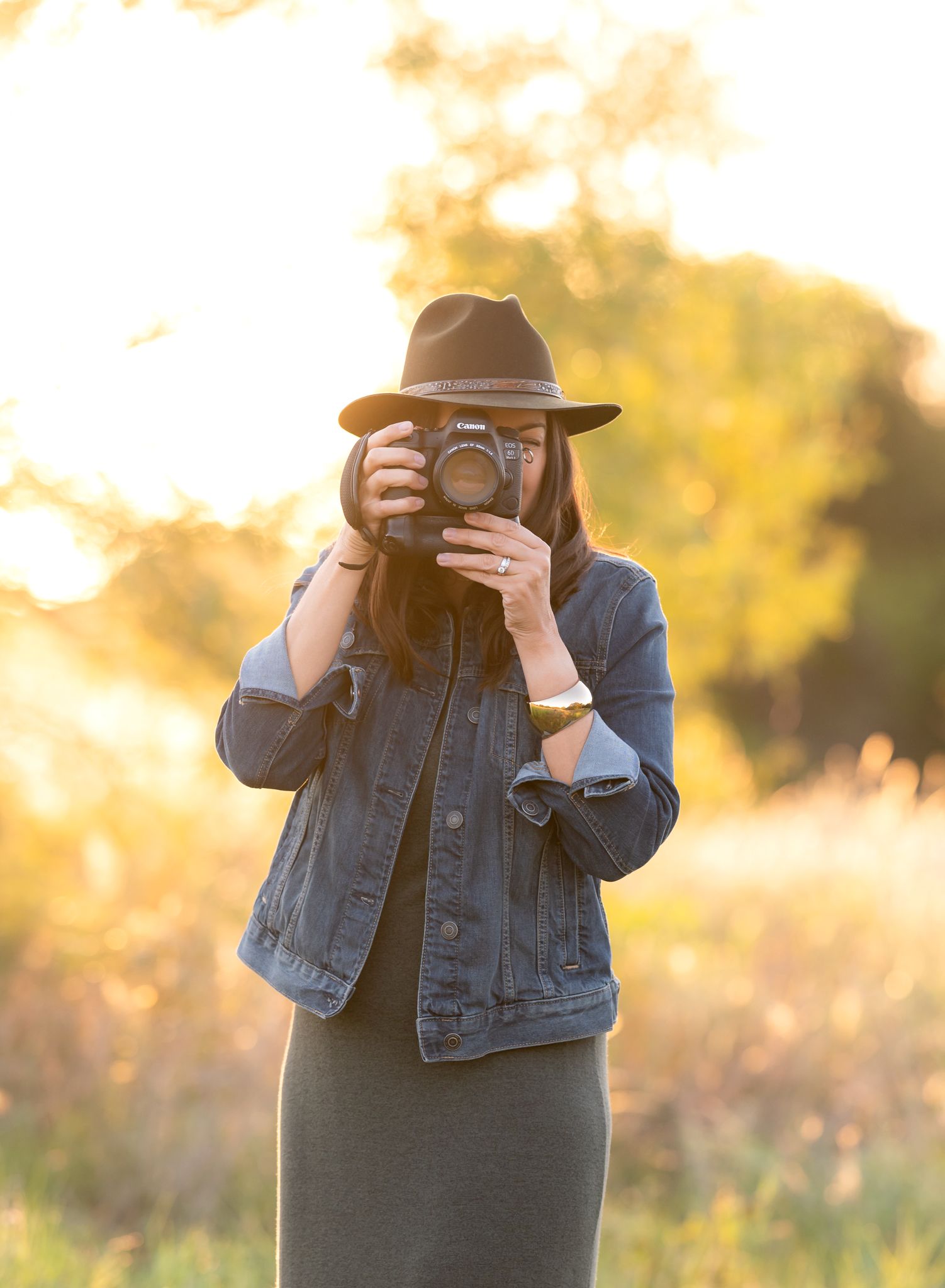 Photographer wearing denim jacket and hat takes photo with vintage camera in golden sunset light.