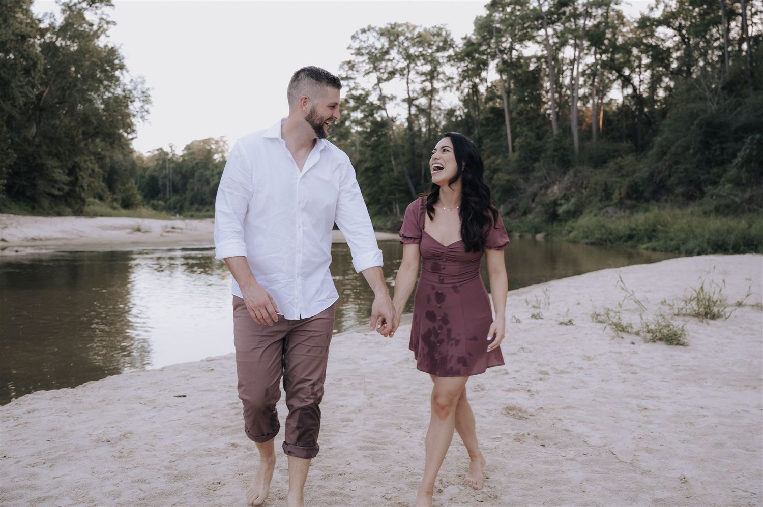 Couple strolling barefoot along sandy riverbank in burgundy dress and white shirt.
