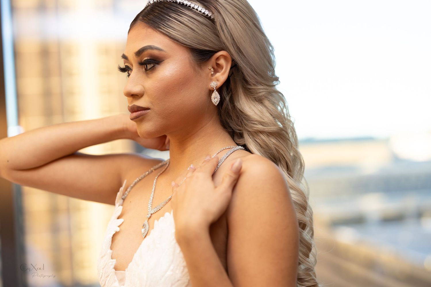 Model with silver blonde hair poses in white dress with elegant jewelry against an urban backdrop during sunset.