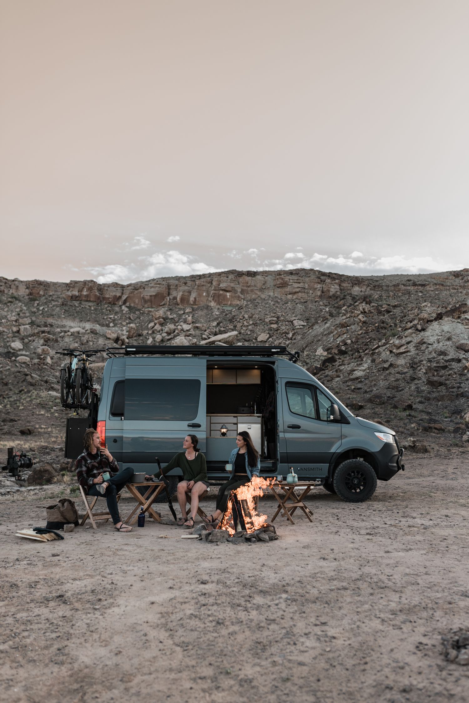 Campsite at dusk with van and campfire in desert landscape.