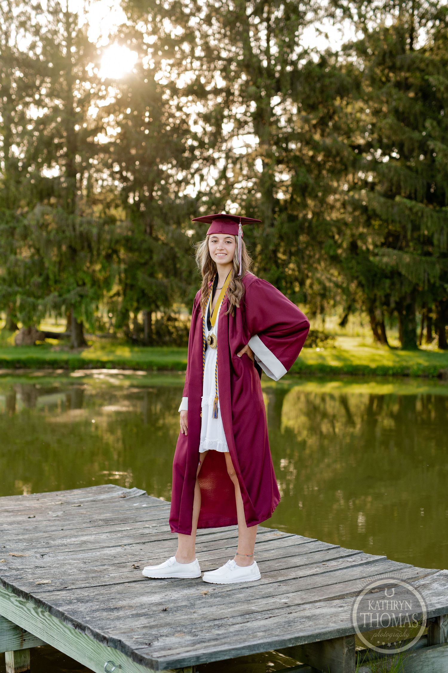 Graduate in burgundy regalia poses on wooden dock by tranquil pond at sunset.