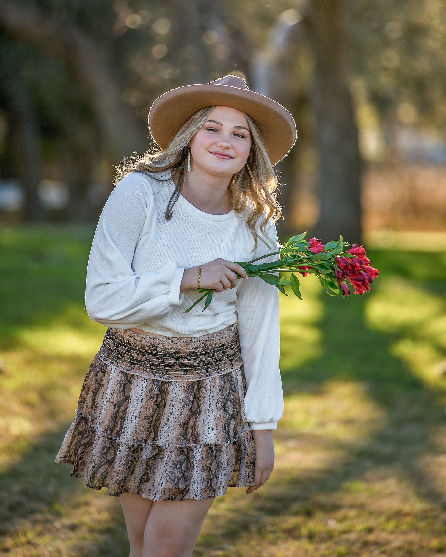 Person in hat and white sweater holding red flowers in sunny outdoor setting.