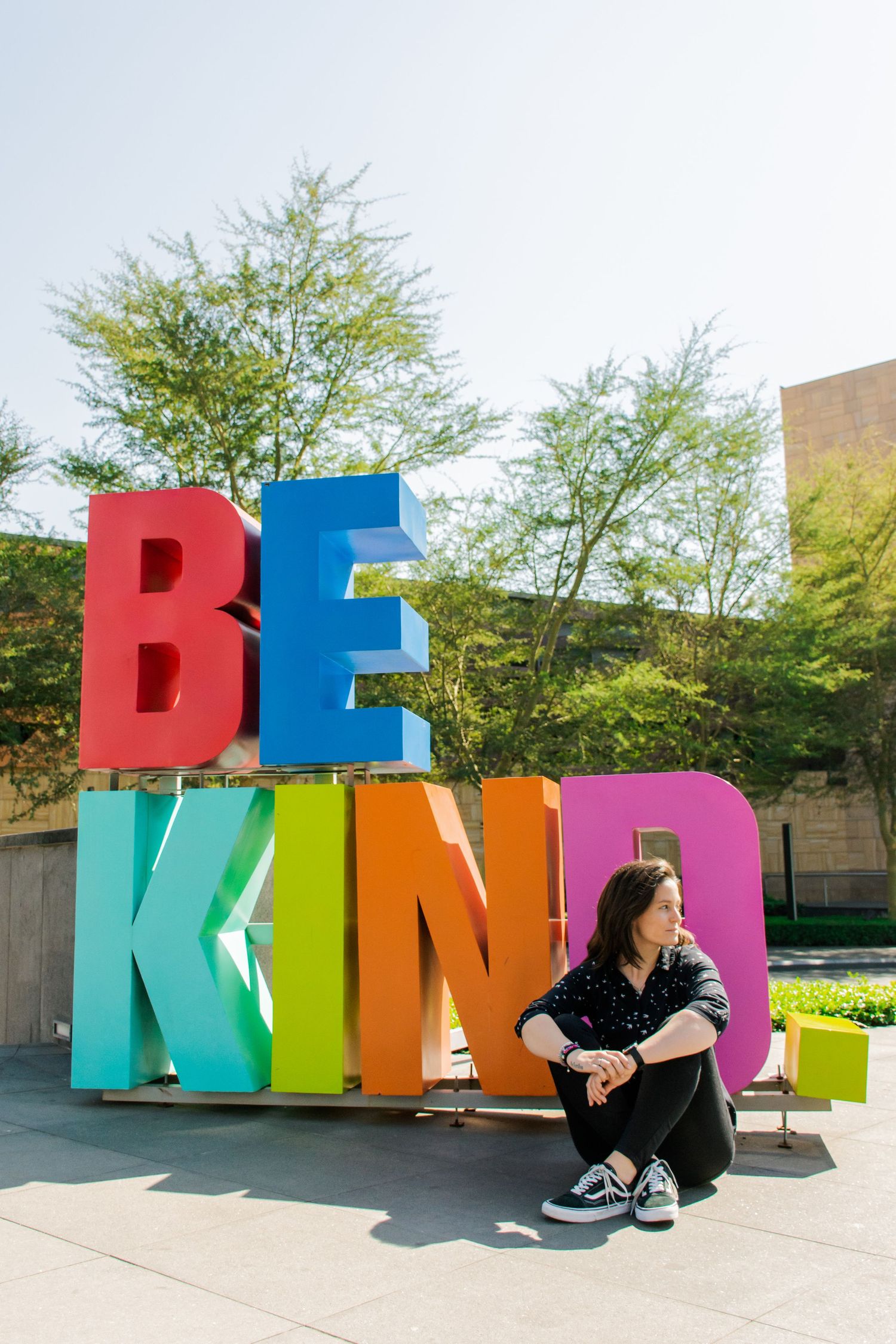 Person sitting next to colorful BE KIND outdoor art installation with trees in background.