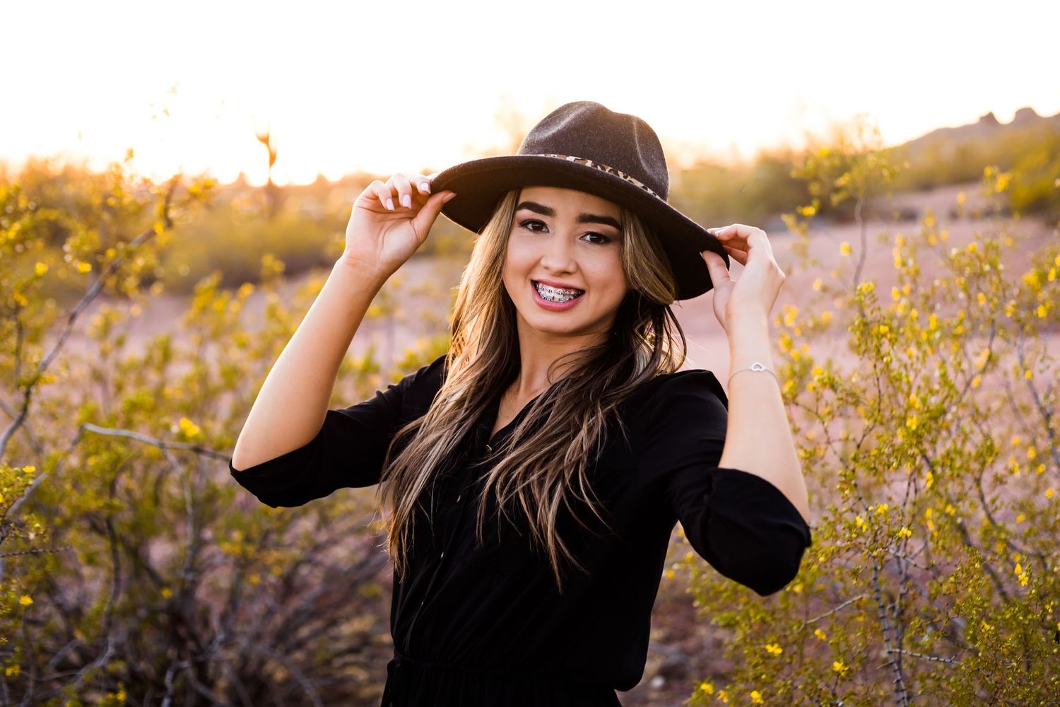 Graduating teenage girl stands in the glowing desert holding her hat in a fashionable way for a smiling senior portrait.