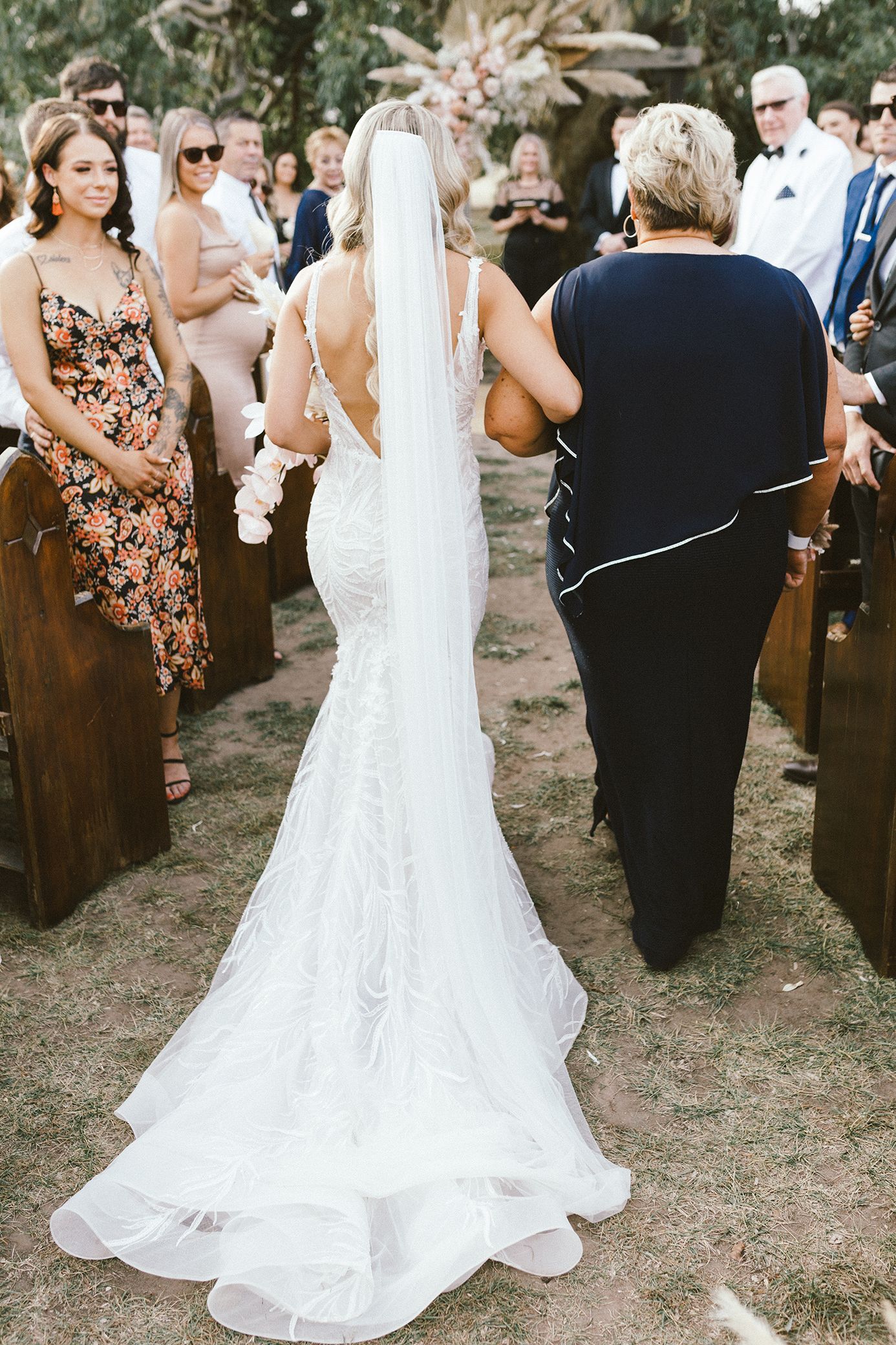 Back view of bride in fitted lace gown with train walking down aisle at outdoor ceremony.