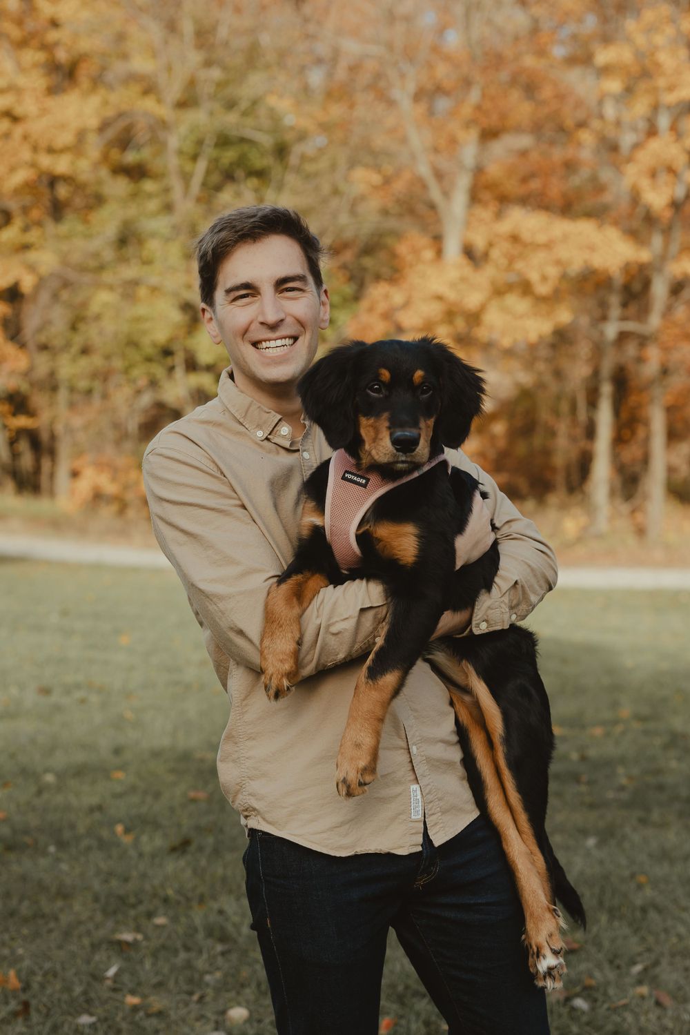 A person in a beige shirt holds a happy black and tan dog in an autumn setting.