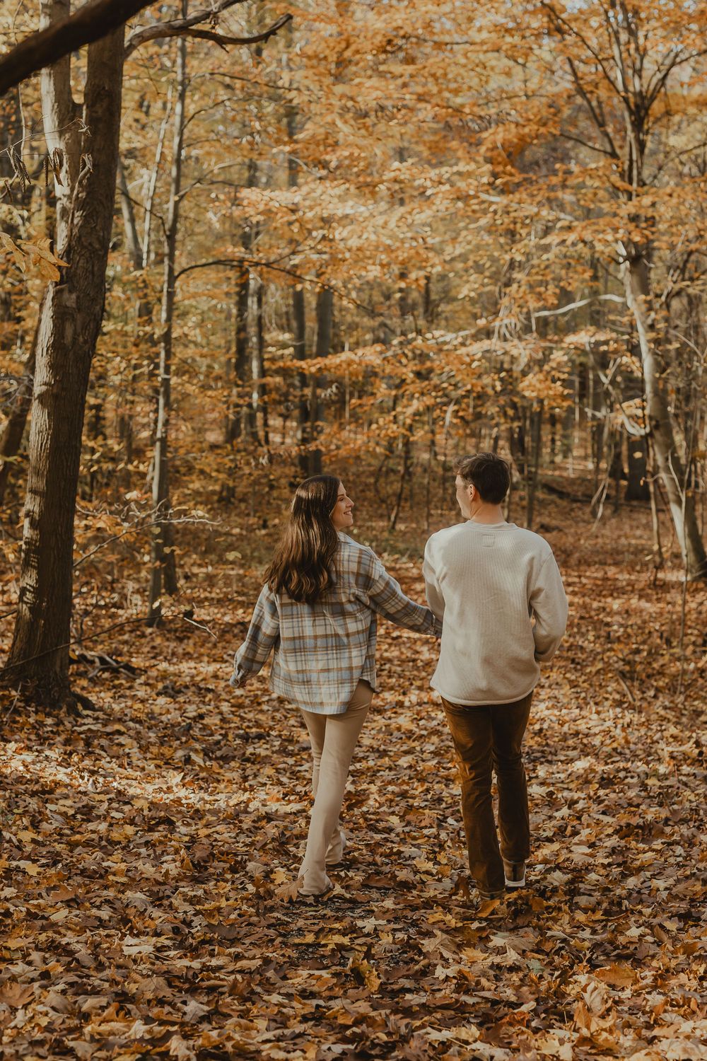A couple walks hand in hand through a forest path covered in golden fall leaves.
