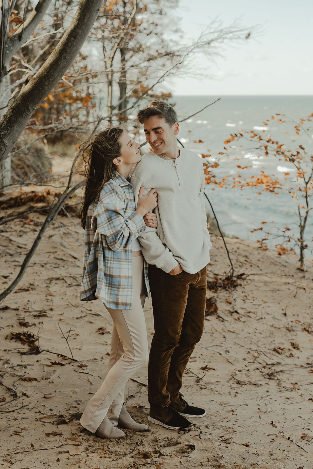 A couple poses lovingly on a beach with autumn trees and water in the background.