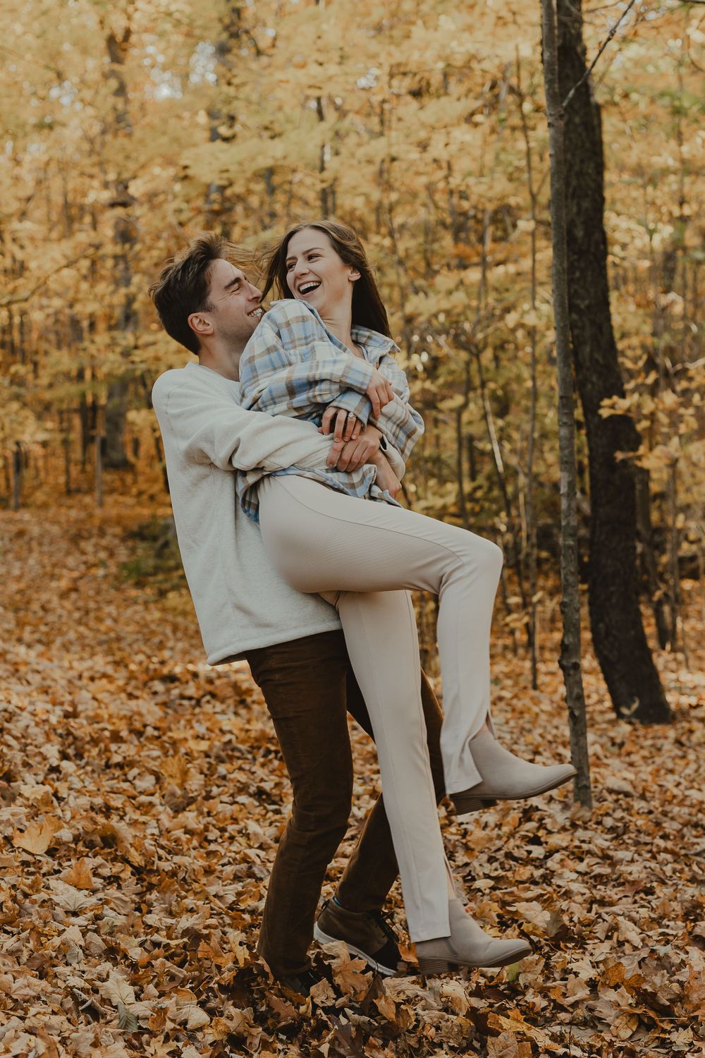 A playful engagement photo of a couple laughing and embracing in an autumn forest setting.