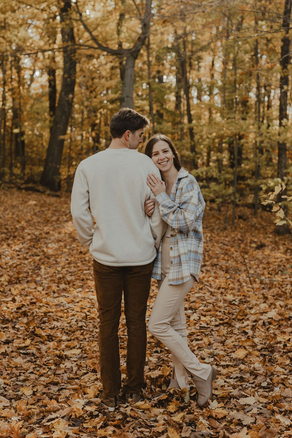 A couple shares a lighthearted moment while standing on fallen leaves in an autumn forest.