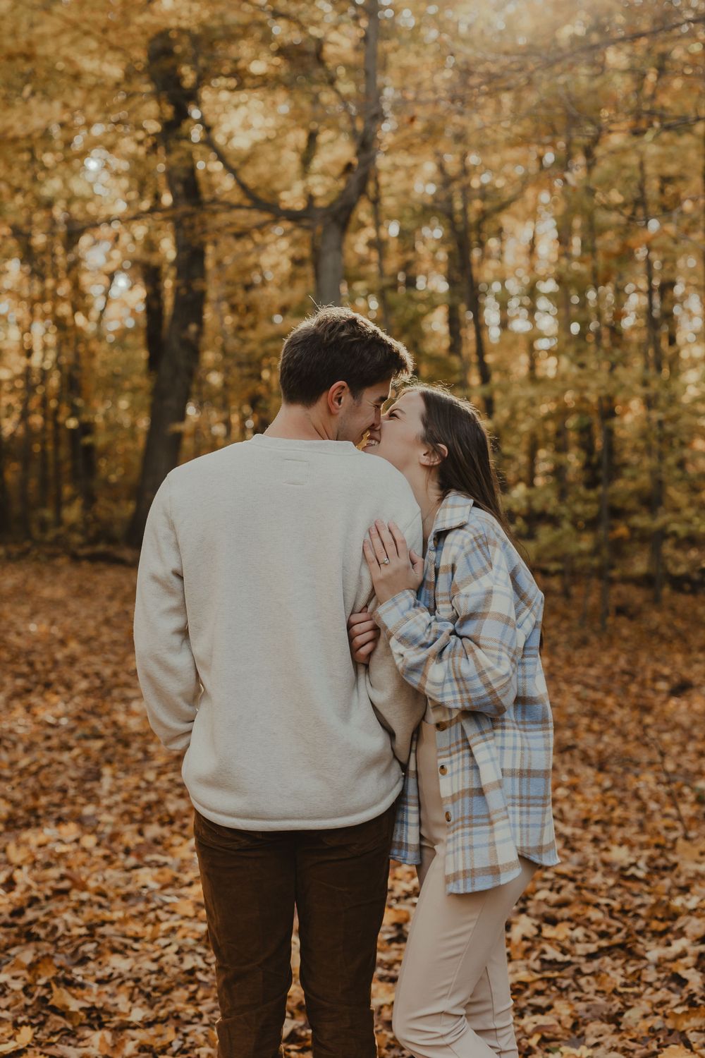 A couple embraces among fallen autumn leaves in a golden-lit forest setting.