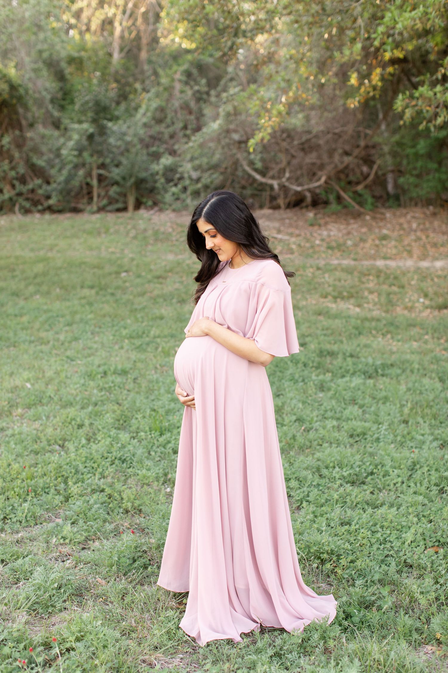 A soft pink maternity gown flows elegantly against a natural wooded backdrop.