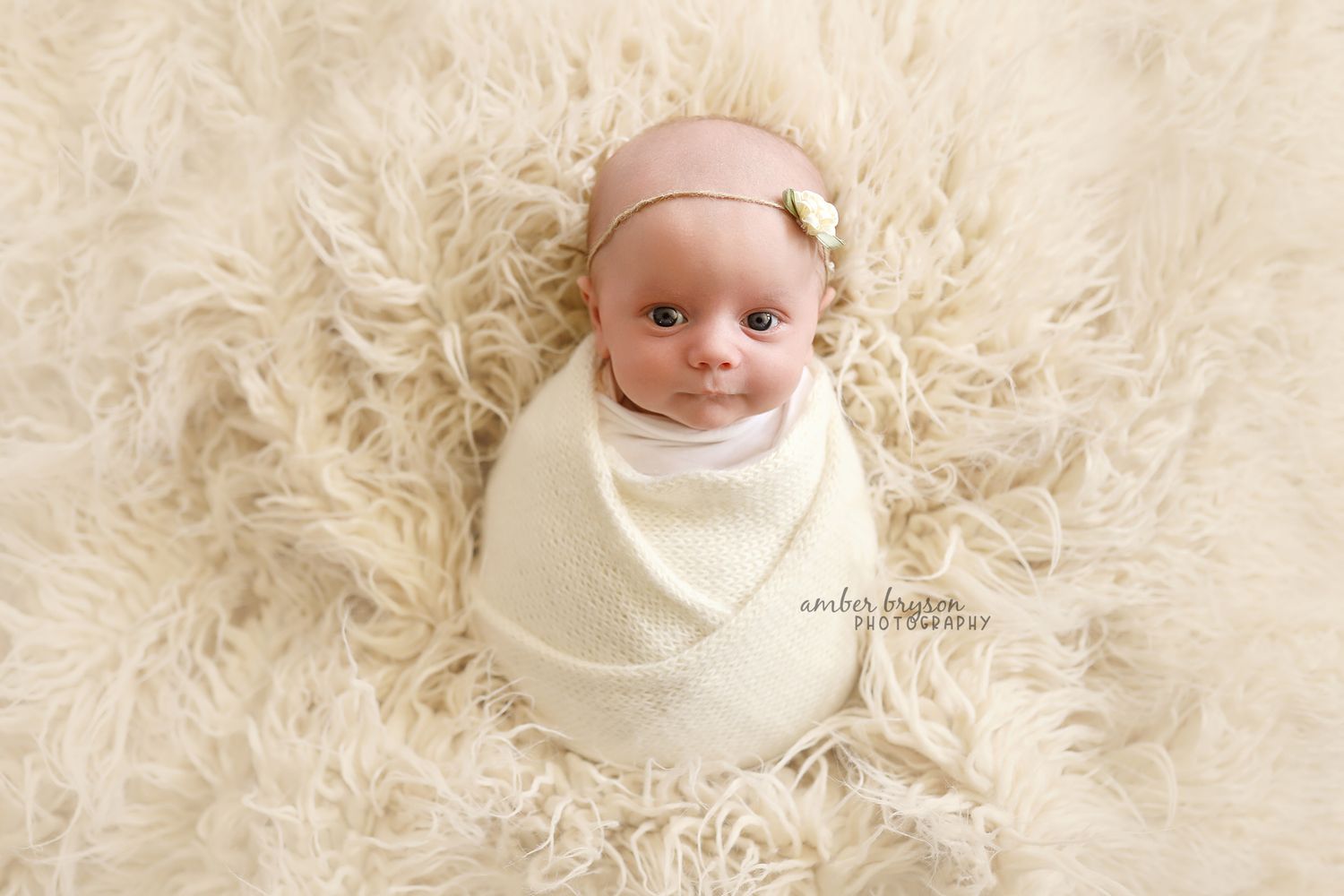 Sleeping newborn with headband wrapped in white on cream fur background.
