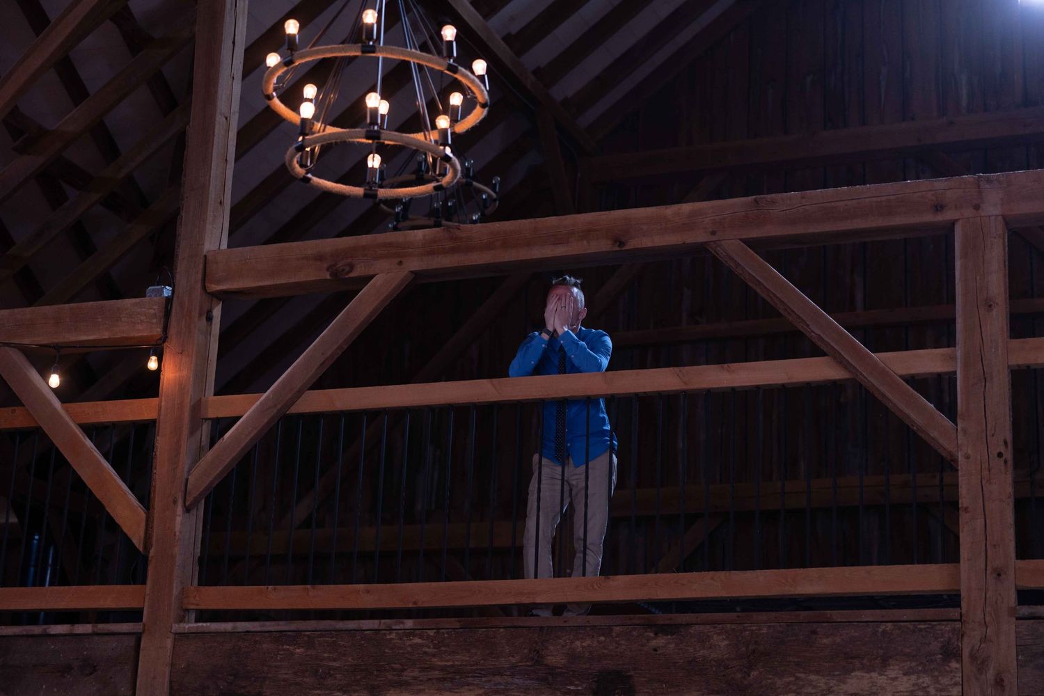 A silhouetted figure stands on a balcony in a barn-style venue with a chandelier visible.