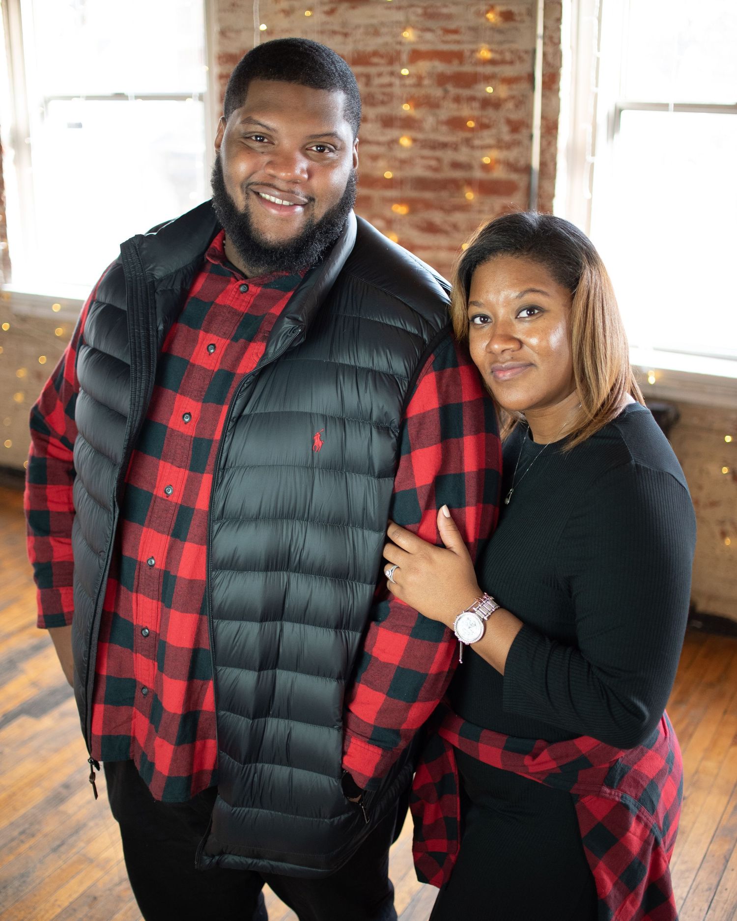 Couple in coordinating buffalo plaid and black outfits pose in brick-walled studio.