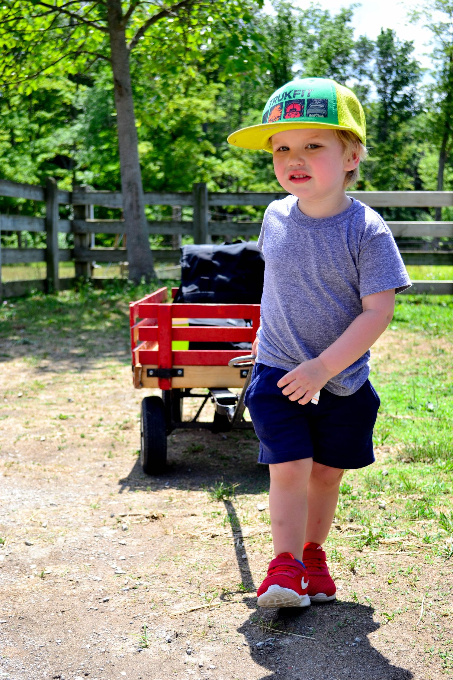 Small child in yellow cap and grey shirt plays near a red wagon in a grassy park.