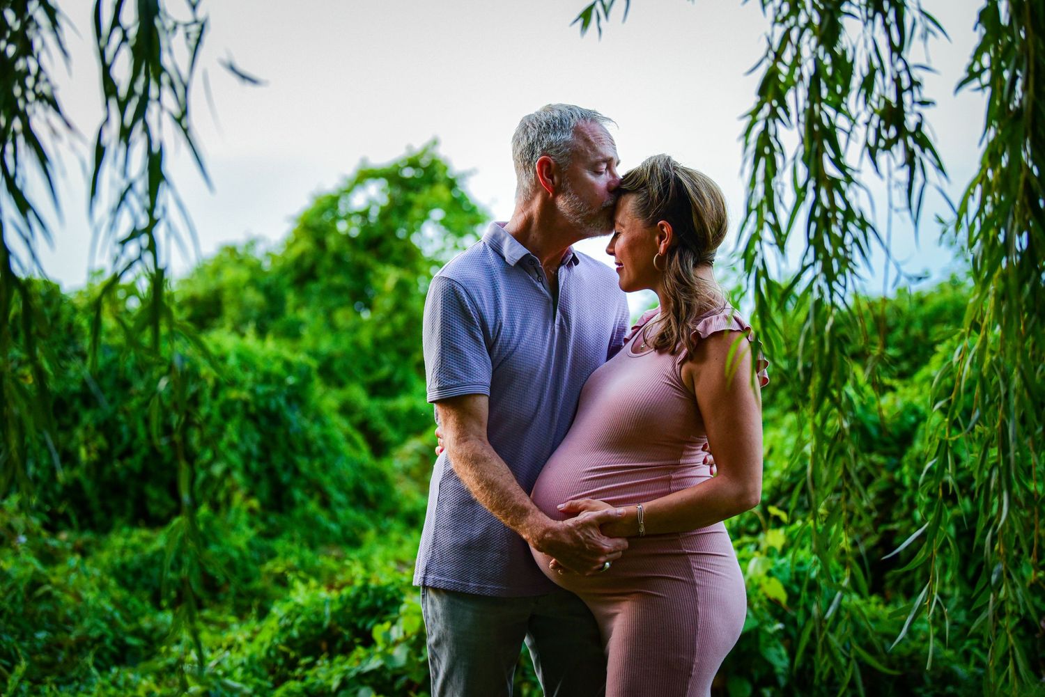 Outdoor maternity photoshoot under willow trees near a tranquil lake.
