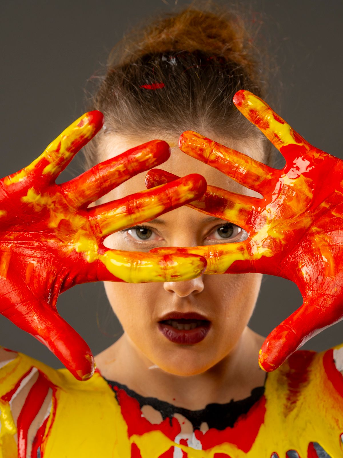 Creative portrait featuring hands painted in bright red and yellow paint held up in front of the subject's eyes.