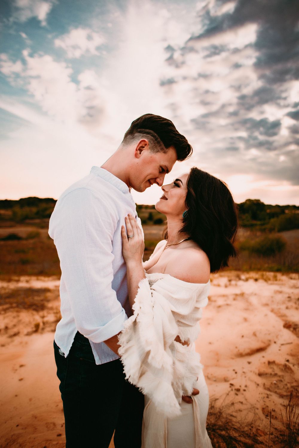 Romantic couple embracing in white attire against dramatic sunset sky.