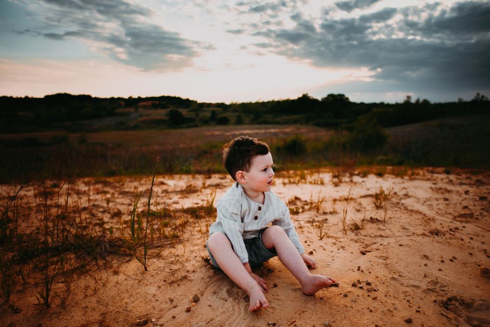 Child sits on sandy ground against dramatic sunset sky and rolling hills in background.