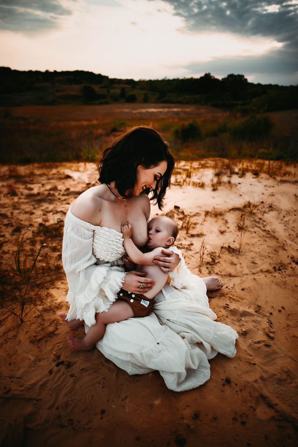 Mother in white dress nurses baby on sandy terrain during golden hour.