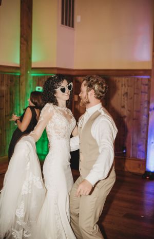 Wedding guests dancing and celebrating with sunglasses in a rustic venue with colored lighting.