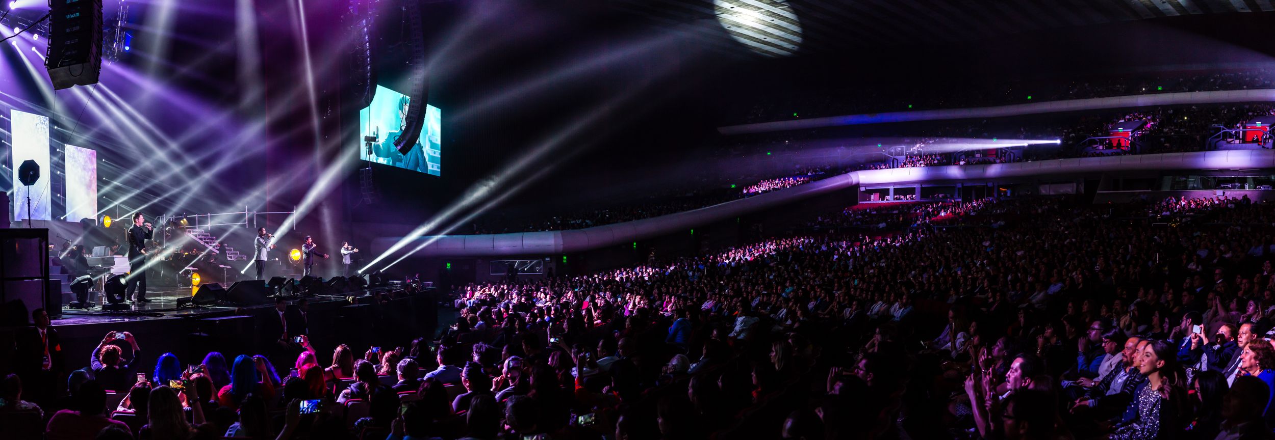 Concert arena filled with audience members under purple stage lighting and beam effects.
