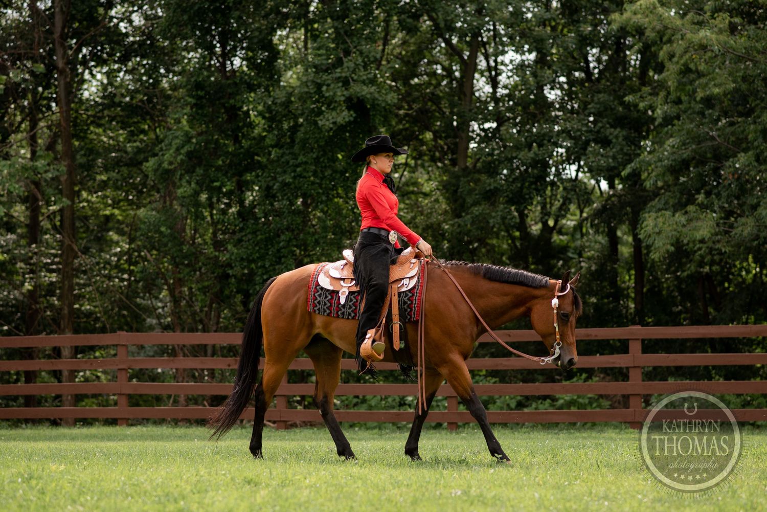 Horse and rider in red shirt riding in fenced arena surrounded by trees.
