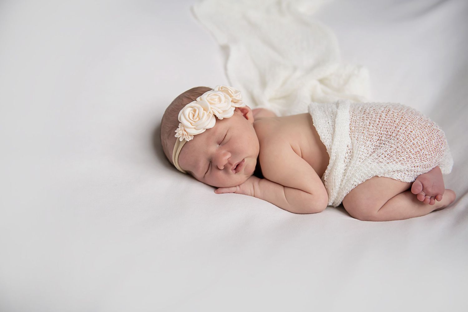 Sleeping newborn in white lace headband and wrap on white backdrop.