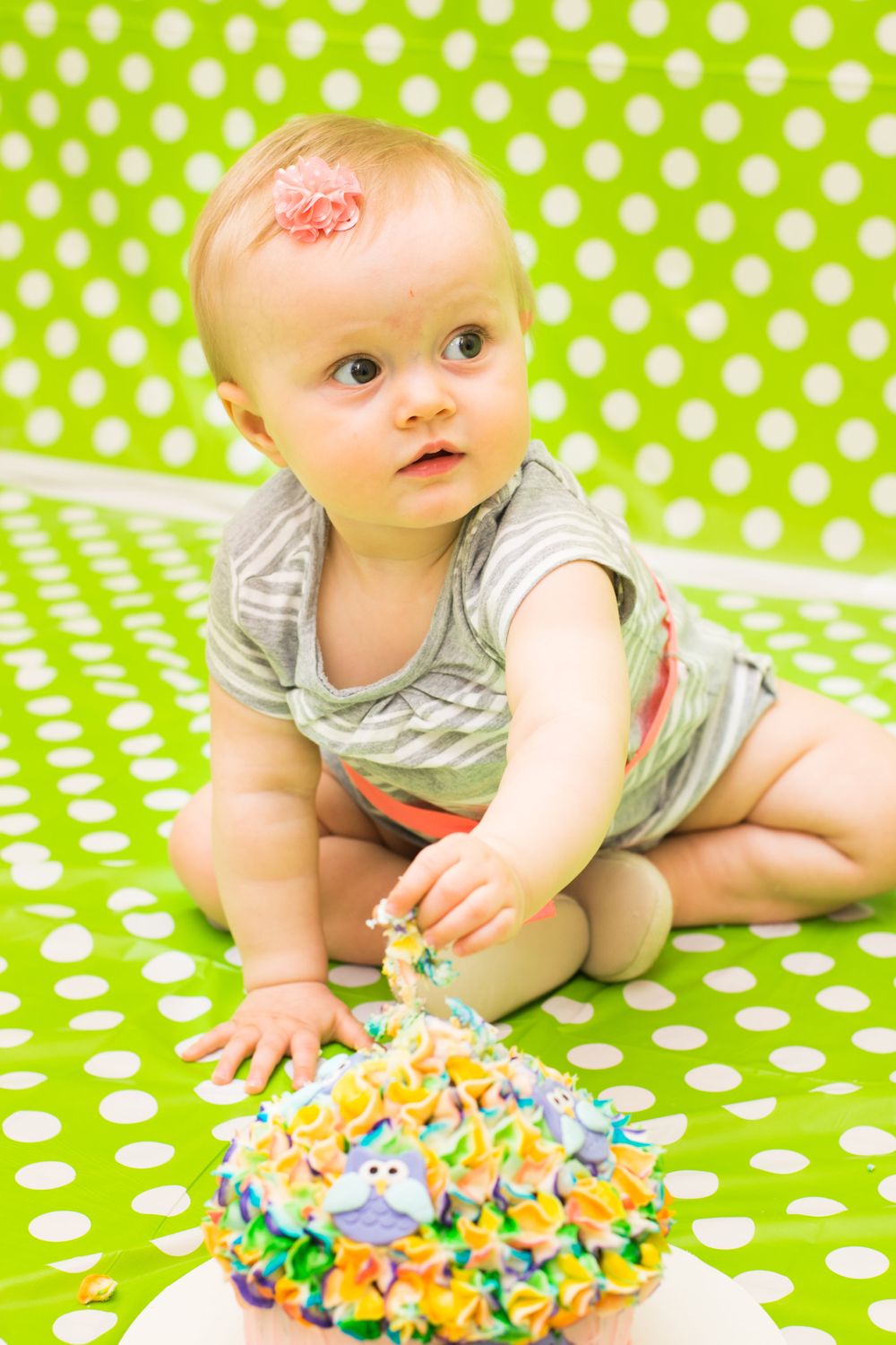 Baby sits on green polka dot backdrop near decorated birthday cake.
