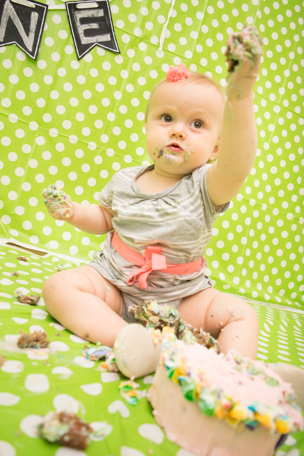 Baby playing with cake during first birthday photoshoot on green polka dot backdrop.