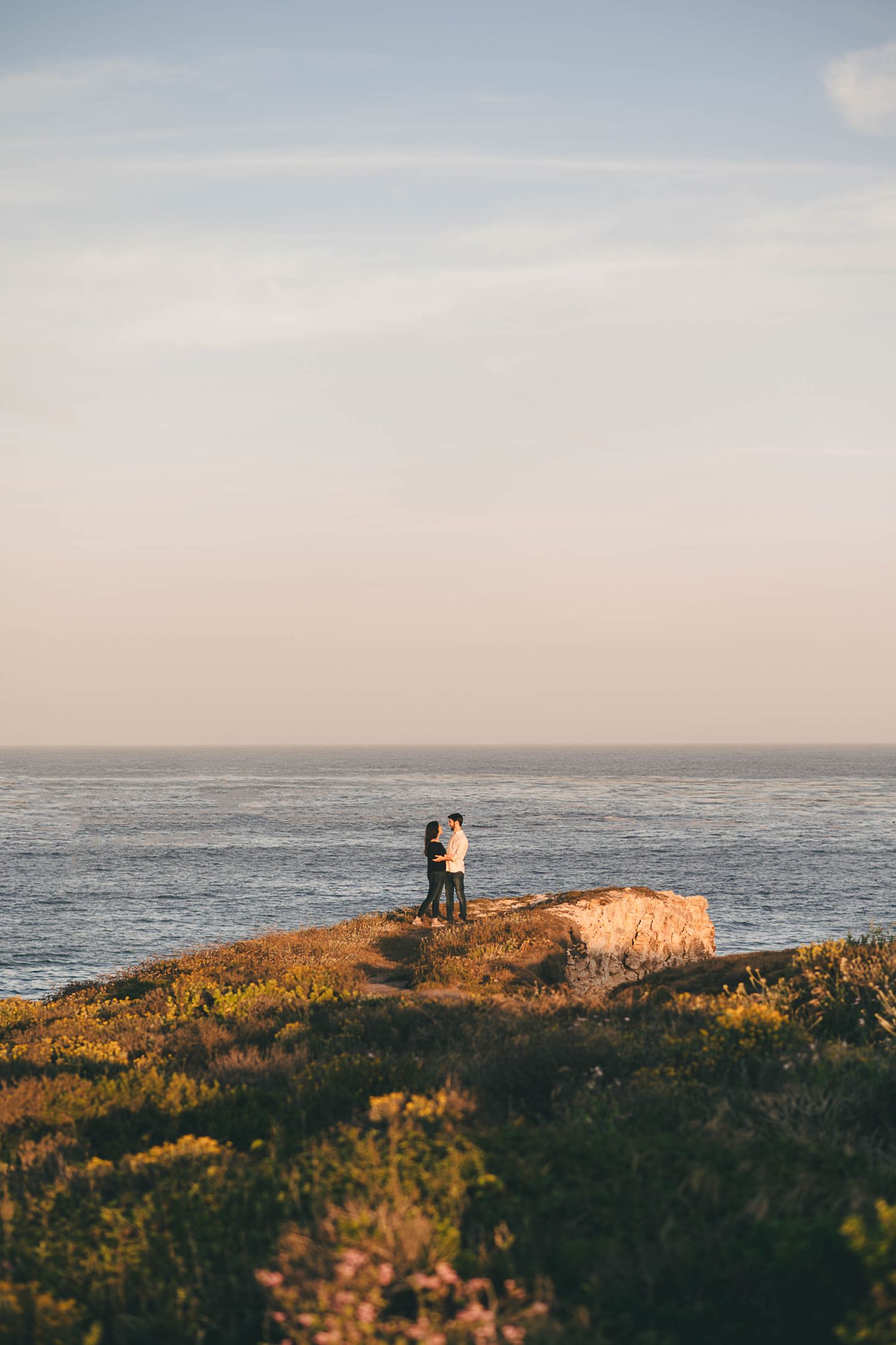 Two silhouettes stand together on a coastal cliff overlooking the ocean at golden hour.